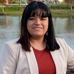 Smiling woman with dark hair and bangs wearing a light blazer and red top, standing by a pond.