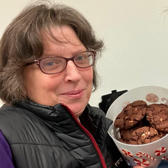 Smiling person wearing glasses and a black vest holding a holiday-themed container filled with chocolate cookies.